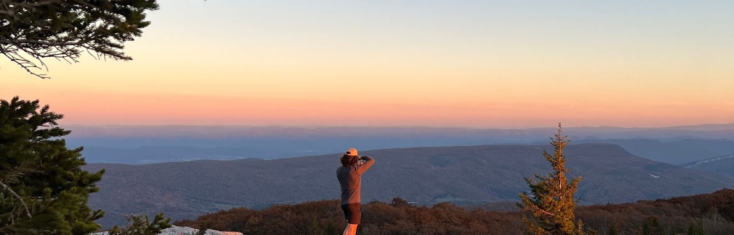 Una persona de pie sobre un afloramiento rocoso con vistas a un paisaje montañoso durante la puesta de sol.