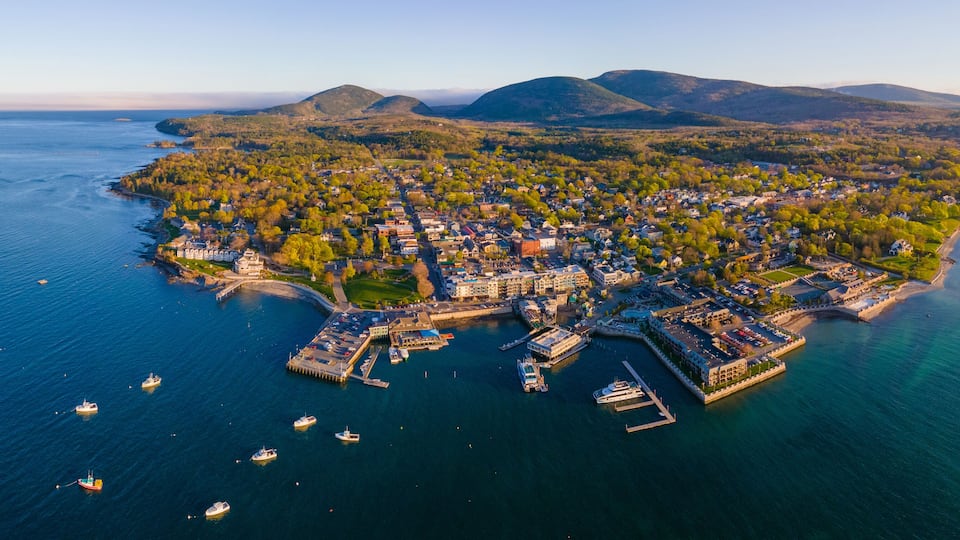 An aerial view of a coastal town with a marina, boats, and surrounding hills.