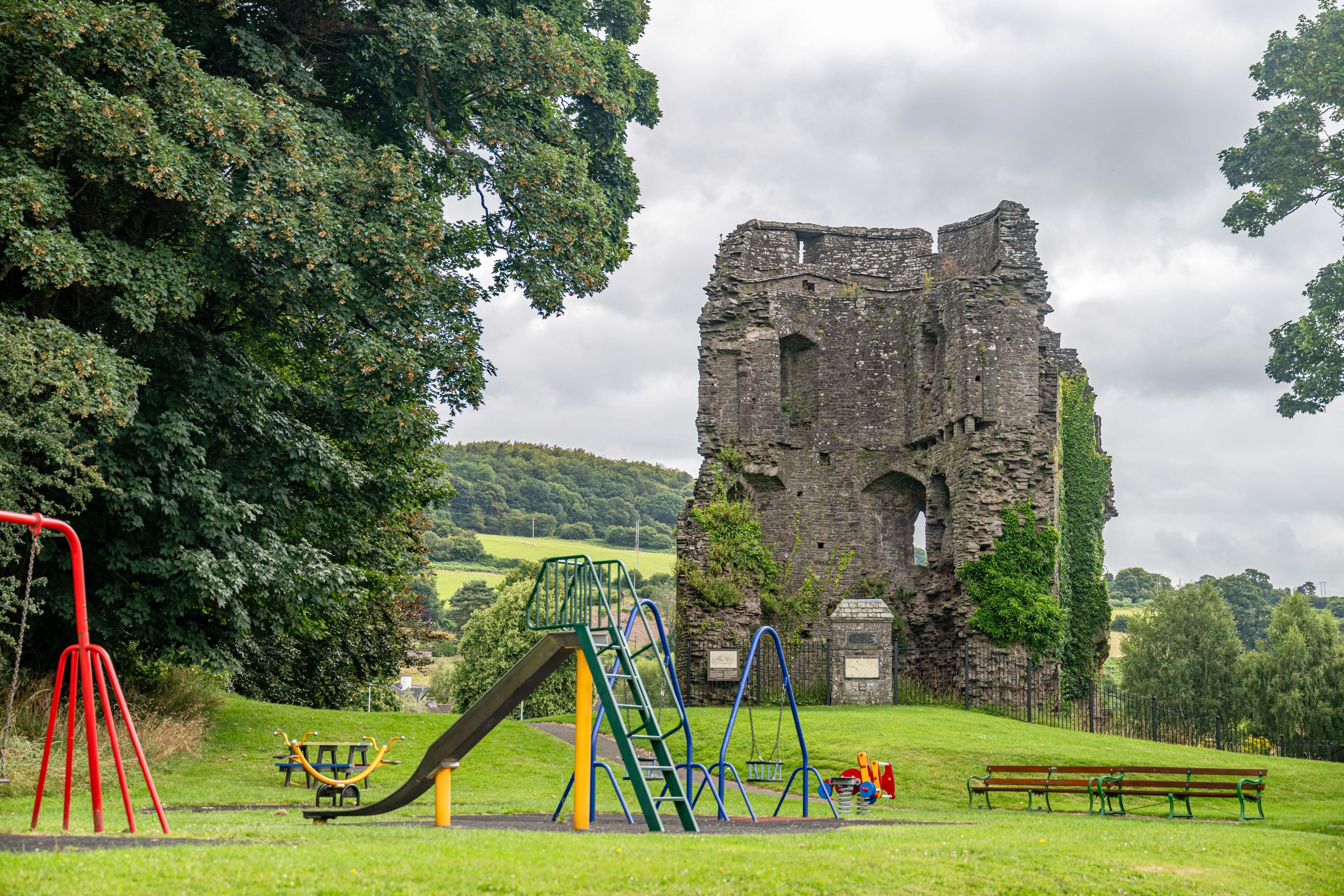 A playground with swings and a slide, a bench, and an ancient stone ruin in the background.