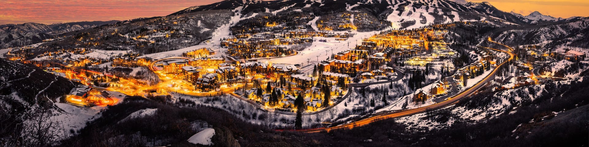A snow-covered mountain town at sunset with illuminated buildings and winding roads.