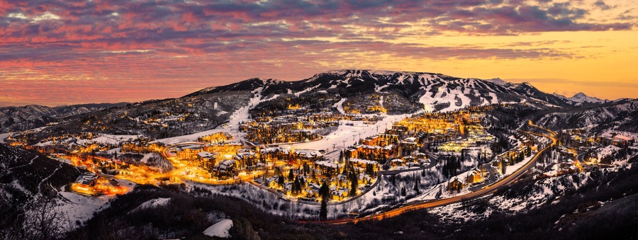 Un pueblo de montaña cubierto de nieve al atardecer, con edificios iluminados y carreteras sinuosas.