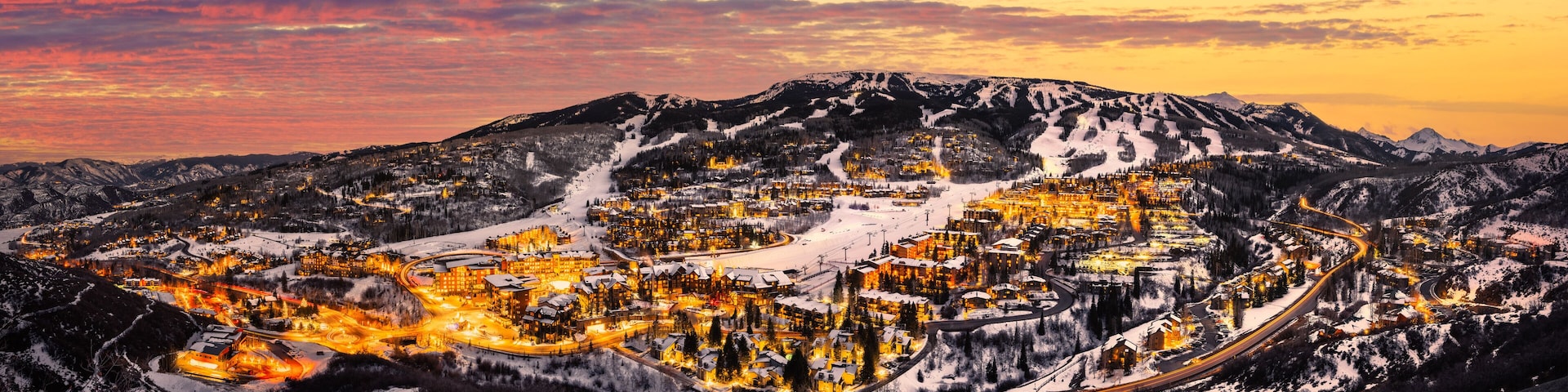Un pueblo de montaña cubierto de nieve al atardecer, con edificios iluminados y carreteras sinuosas.
