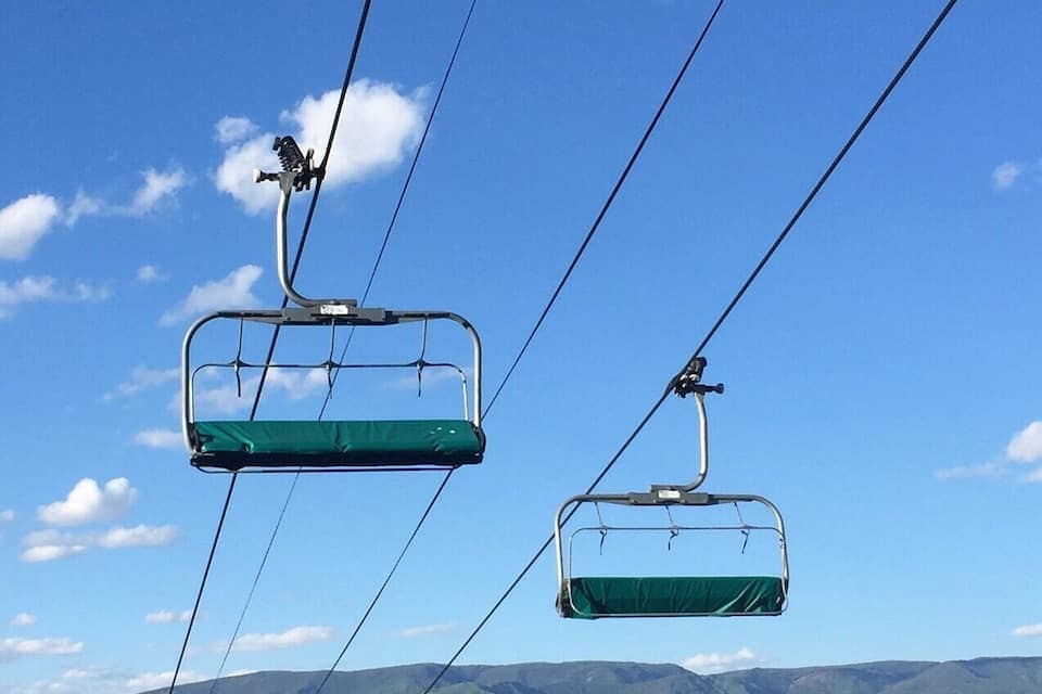 Um teleférico com cadeiras suspensas por cabos contra um céu azul límpido, com vista para uma paisagem montanhosa e uma aldeia abaixo.