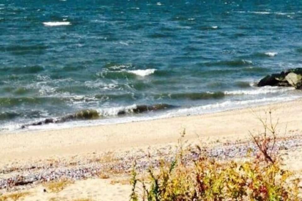 A beach with waves, a sailboat, and a distant city skyline.