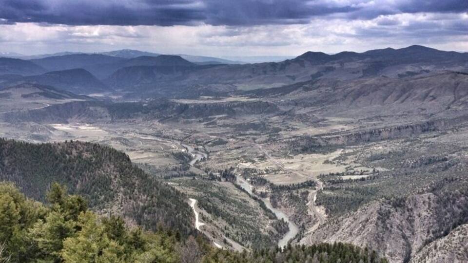 A mountainous landscape with a winding river, dense forests, and distant hills under a cloudy sky.