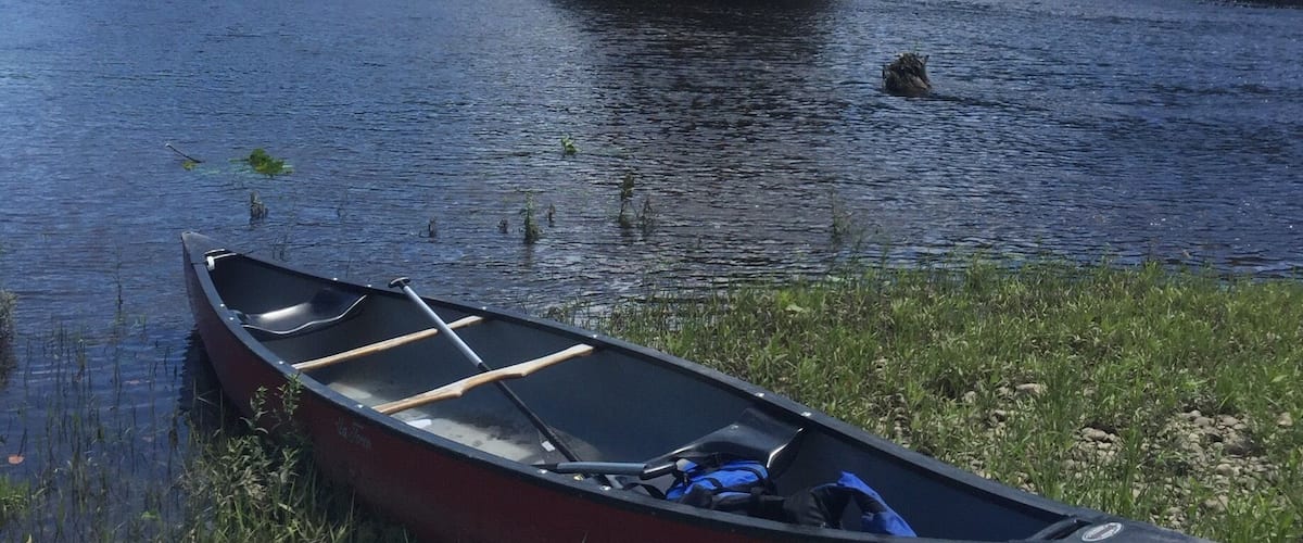 A red canoe on a grassy bank by a calm river, with a rocky outcrop in the background.