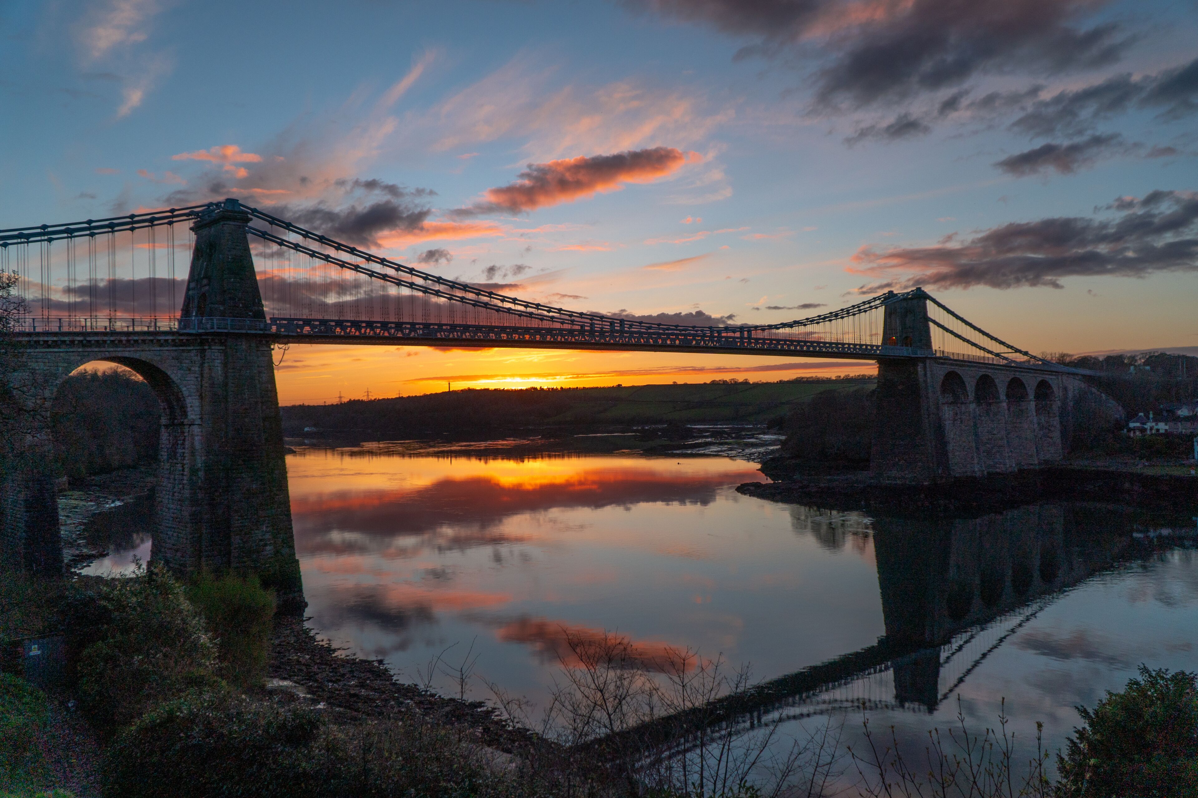 Uma ponte suspensa sobre um rio calmo ao pôr do sol, com céu limpo.
