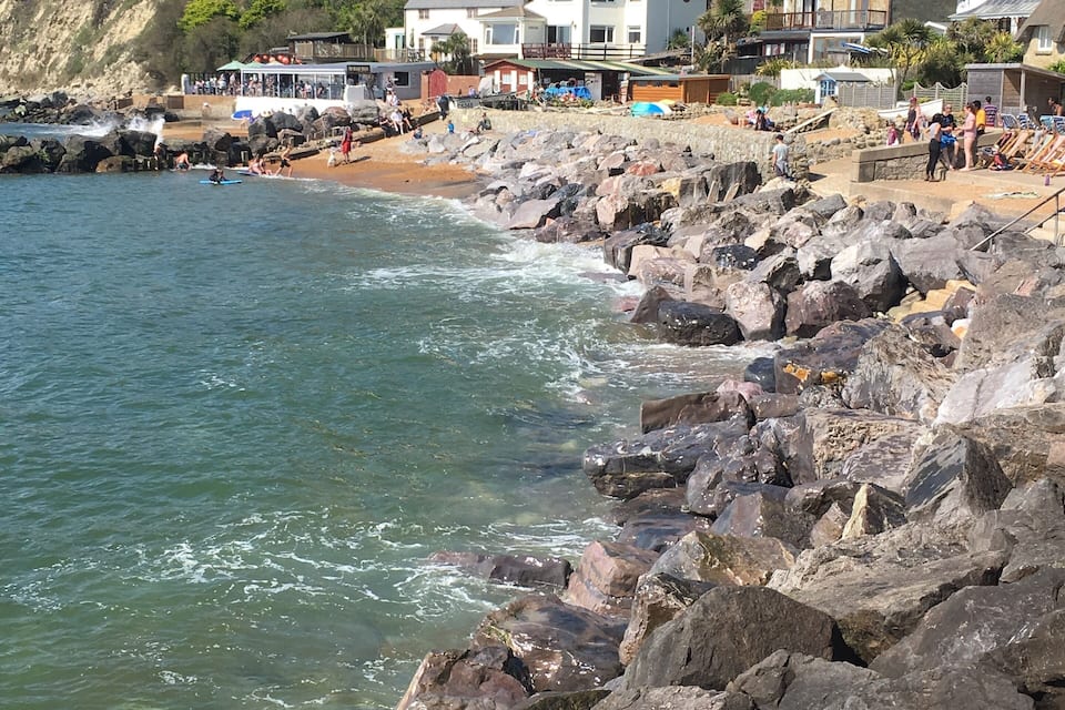 Una costa rocosa con olas rompiendo en la orilla, una playa de arena y edificios al fondo.