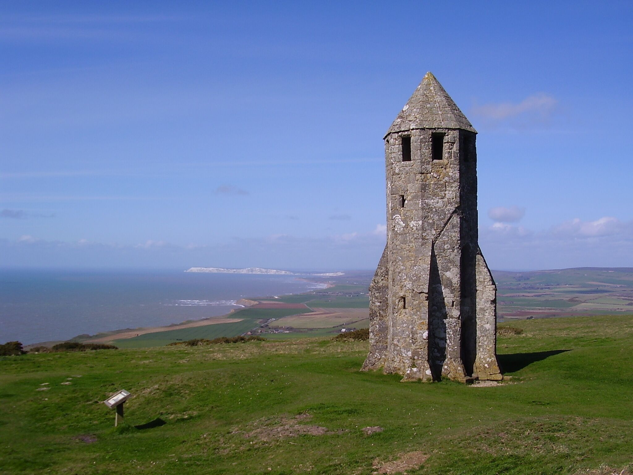 A stone tower with a conical roof on a grassy hill overlooking a coastal landscape.