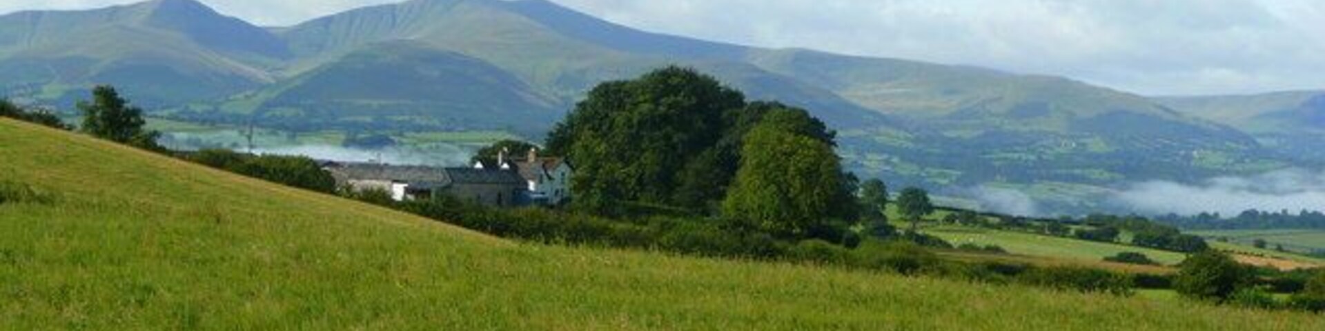A grassy field with a house and trees in the foreground, and rolling hills in the background.