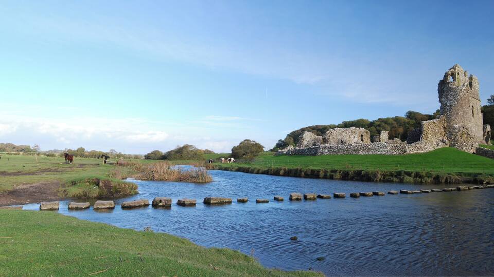 A historic stone tower, a river with stepping stones, and grazing horses in a rural landscape.