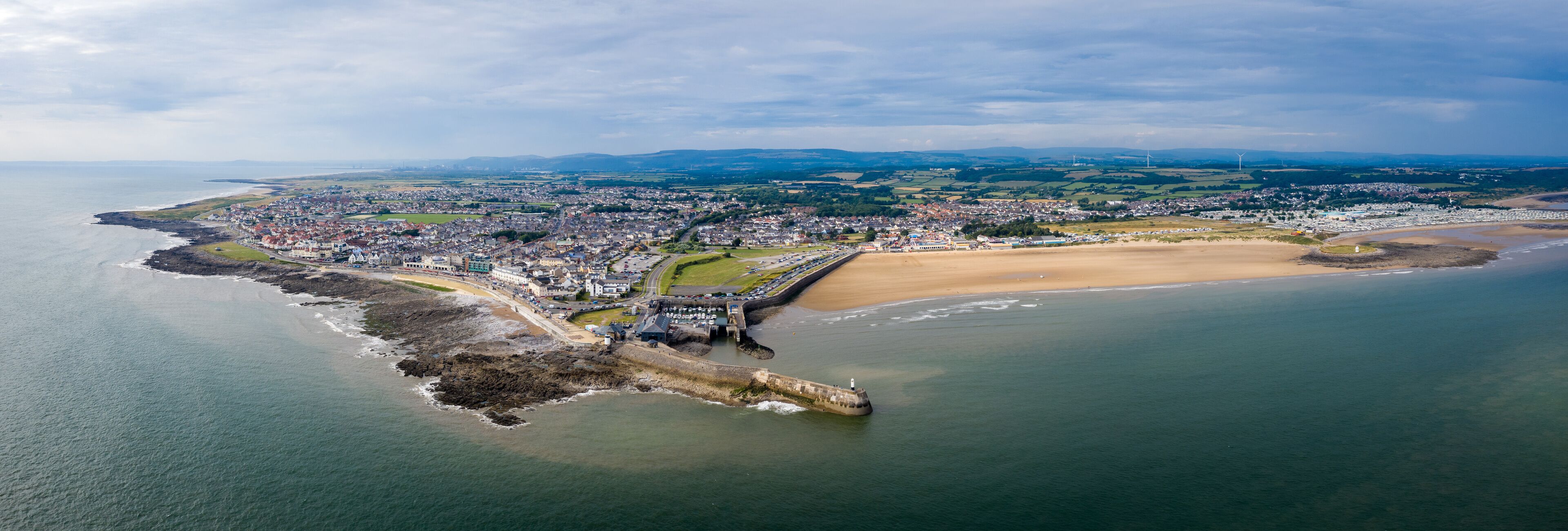 An aerial view of a coastal town with a prominent breakwater, sandy beach, and rolling hills in the background.