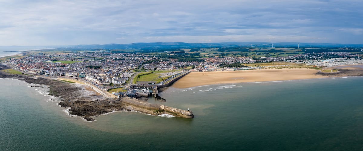 An aerial view of a coastal town with a prominent breakwater, sandy beach, and rolling hills in the background.