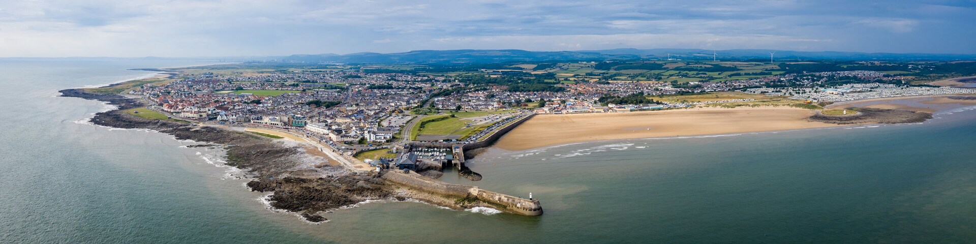 An aerial view of a coastal town with a prominent breakwater, sandy beach, and rolling hills in the background.
