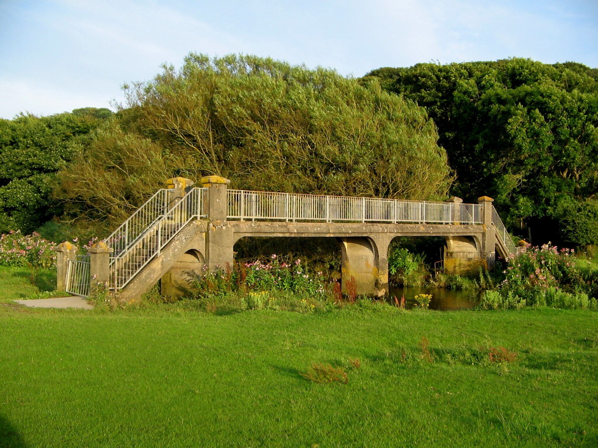 A stone bridge with a metal railing over a river, surrounded by lush greenery and flowering plants.