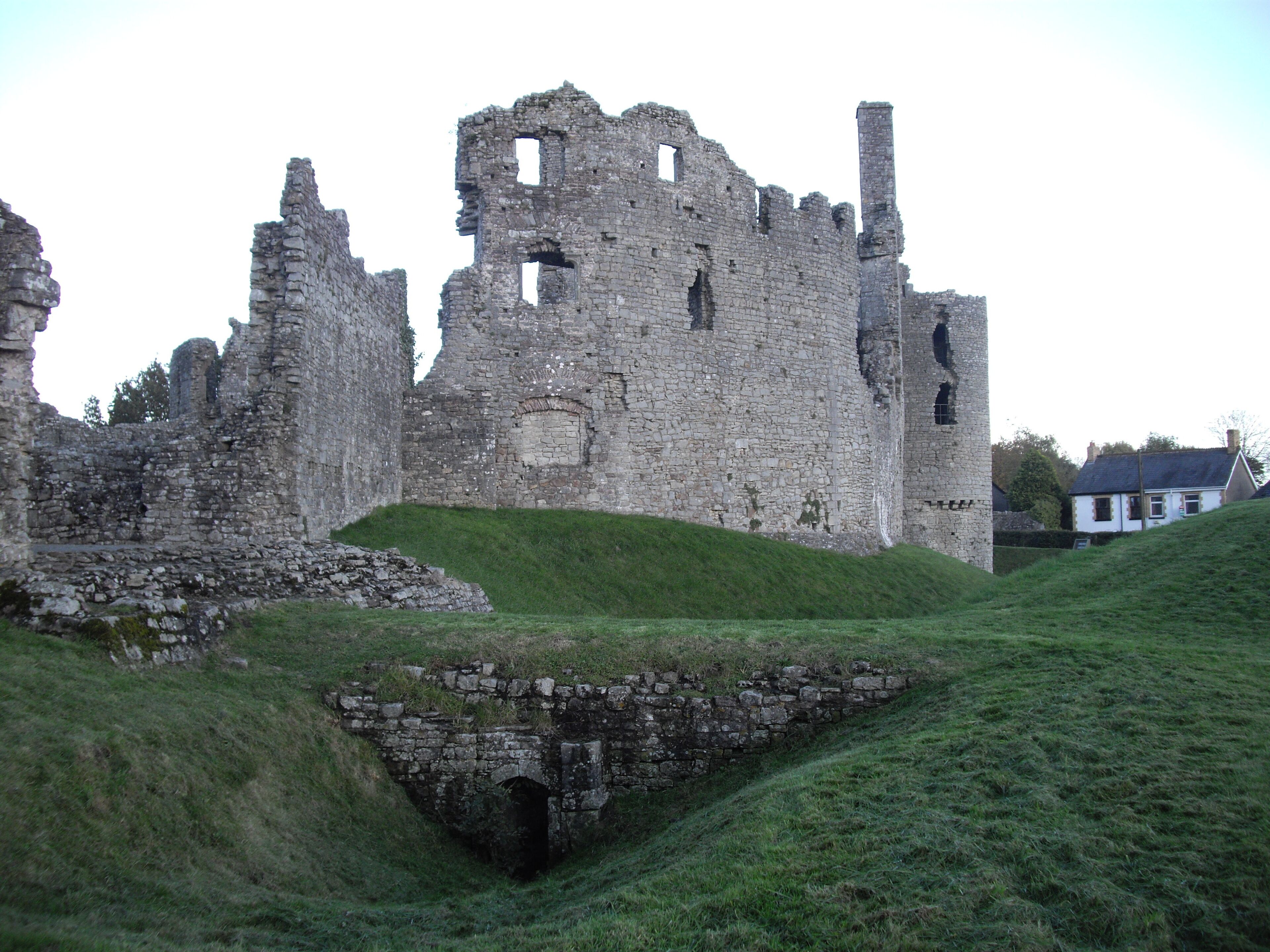 Ruinas de un castillo de piedra histórica con una colina cubierta de hierba y una casa blanca al fondo.