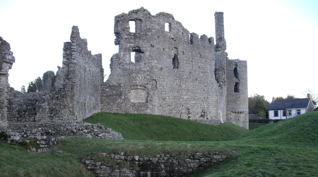 Ruinas de un castillo de piedra histórica con una colina cubierta de hierba y una casa blanca al fondo.