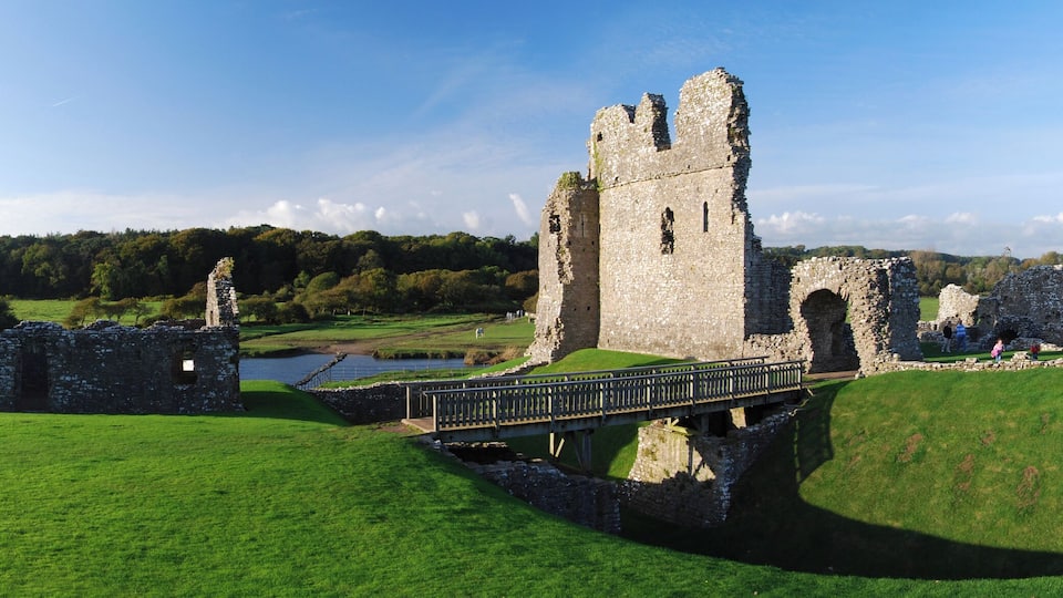 A historic castle ruin with a bridge, surrounded by lush green fields and a river.