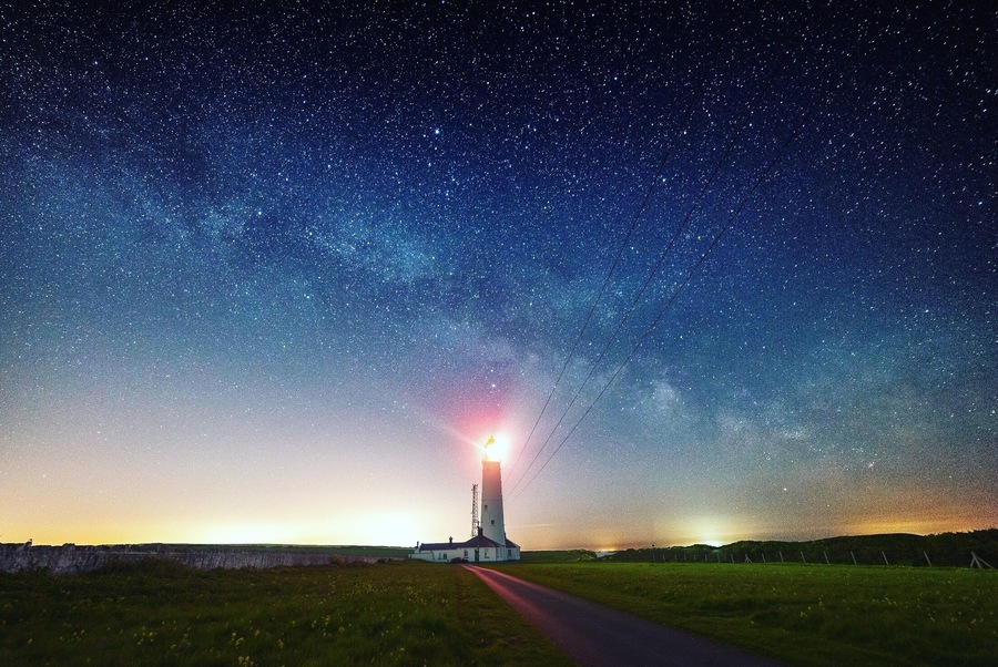 Um farol à noite sob um céu estrelado, com um caminho visível que o leva até ele.