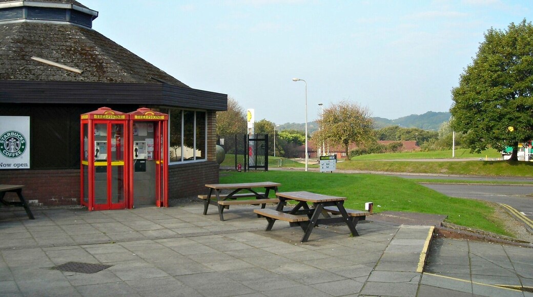 Una cabina telefónica roja, un letrero de Starbucks y mesas de picnic sobre una superficie pavimentada, con un campo cubierto de césped y colinas al fondo.
