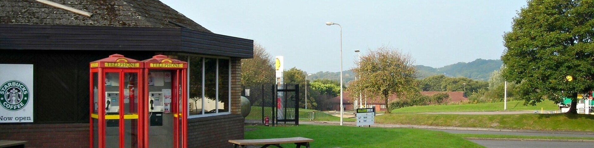 A red telephone booth, a Starbucks sign, and picnic tables on a paved area with a grassy field and hills in the background.