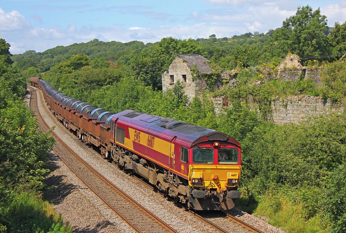 Um trem vermelho e amarelo percorre uma paisagem verdejante com uma estrutura de pedra ao fundo.