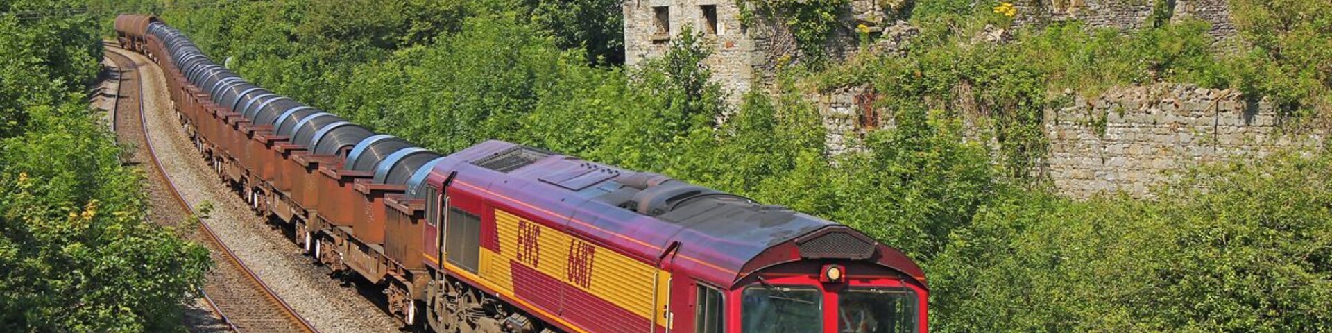 Um trem vermelho e amarelo percorre uma paisagem verdejante com uma estrutura de pedra ao fundo.