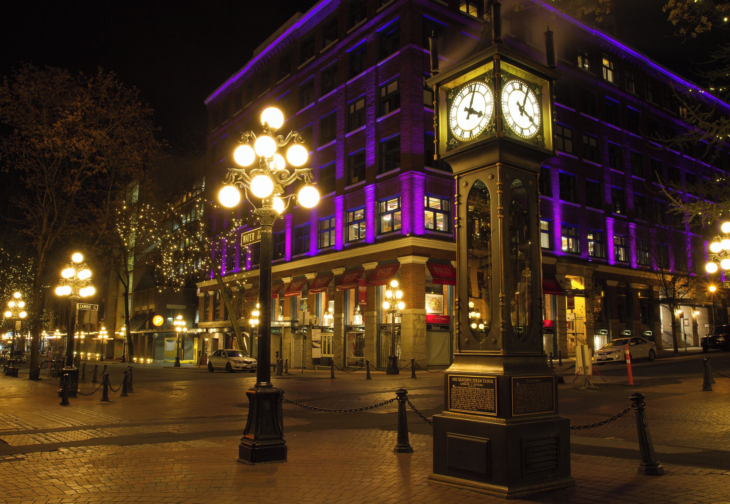 A historic building with a prominent clock tower, illuminated by purple lights, and a well-lit street with decorative street lamps.