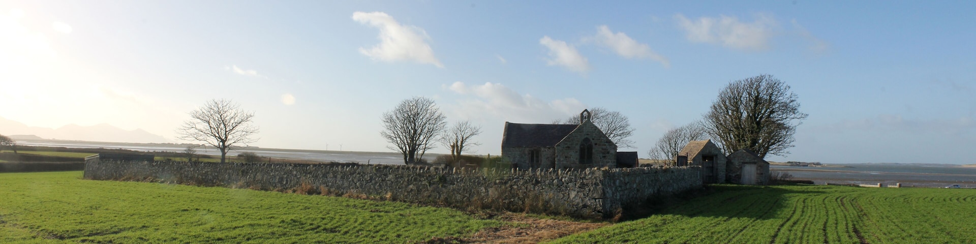 A rural landscape with a stone wall, a stone building, and a field of crops.
