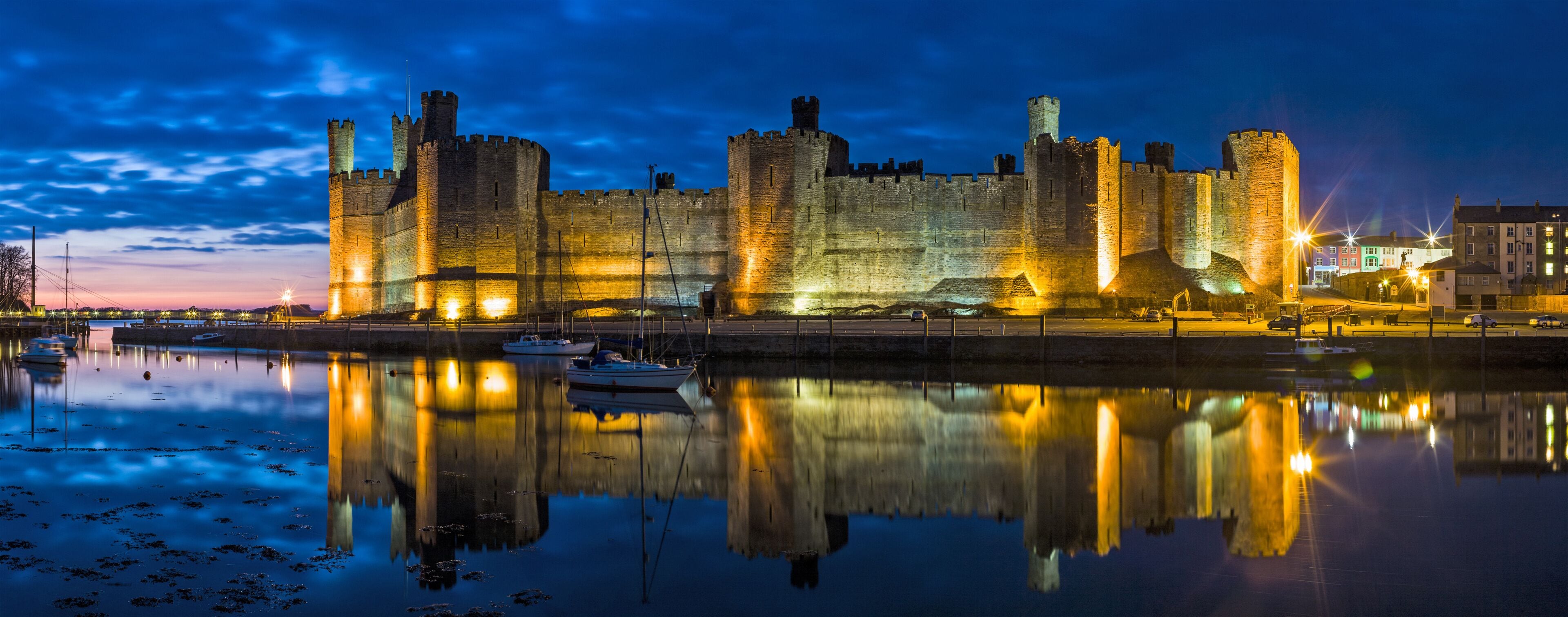 Um castelo histórico com várias torres e um fosso, refletindo na água calma.