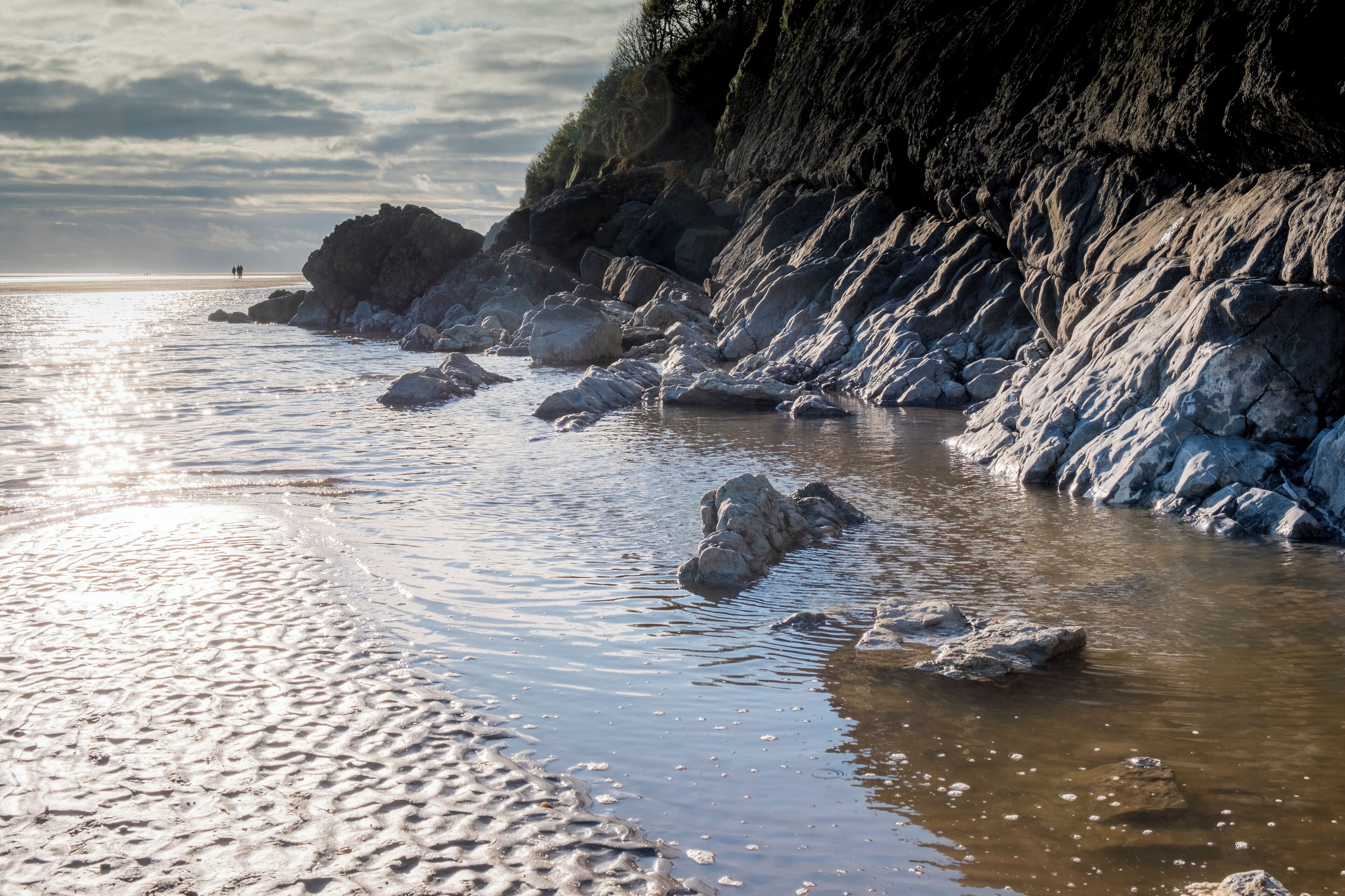 A rocky shoreline with waves gently lapping against the rocks and a distant view of people on the beach.
