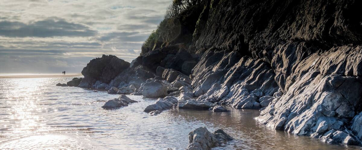A rocky shoreline with waves gently lapping against the rocks and a distant view of people on the beach.