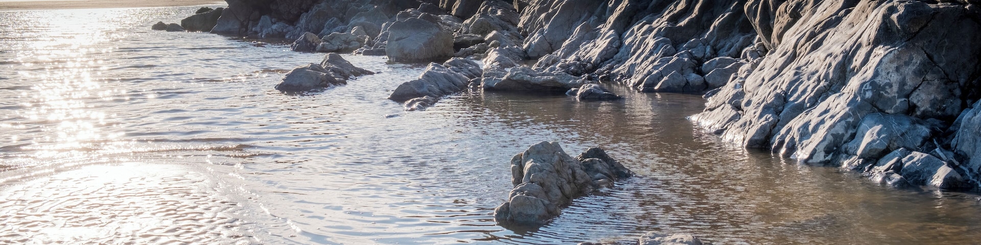 A rocky shoreline with waves gently lapping against the rocks and a distant view of people on the beach.