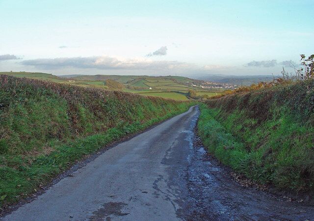 A narrow rural road flanked by grassy fields and rolling hills.