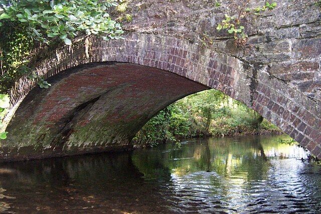 A stone bridge arching over a calm river, surrounded by greenery.