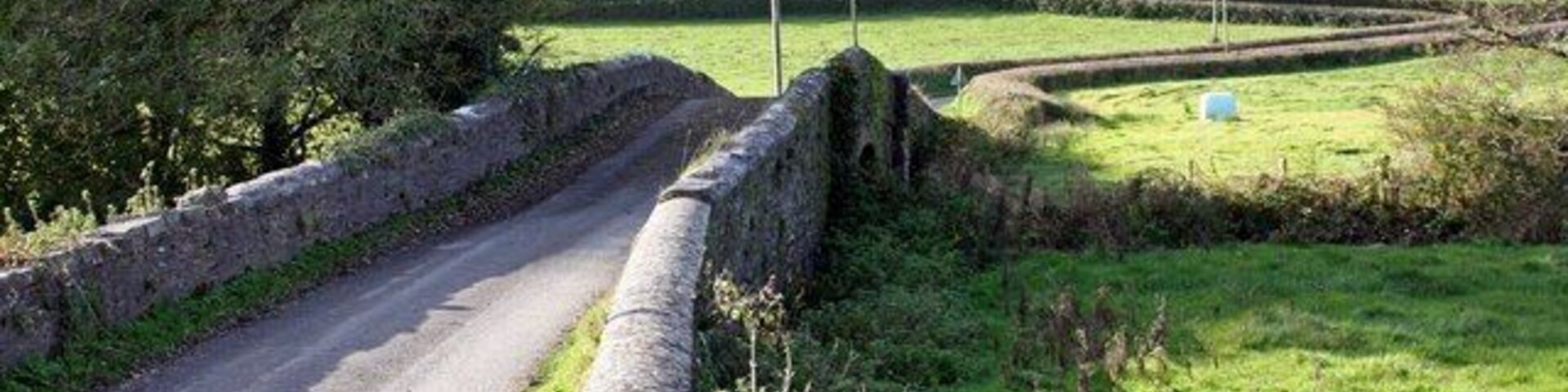 A narrow rural road bordered by stone walls, surrounded by green fields and trees.