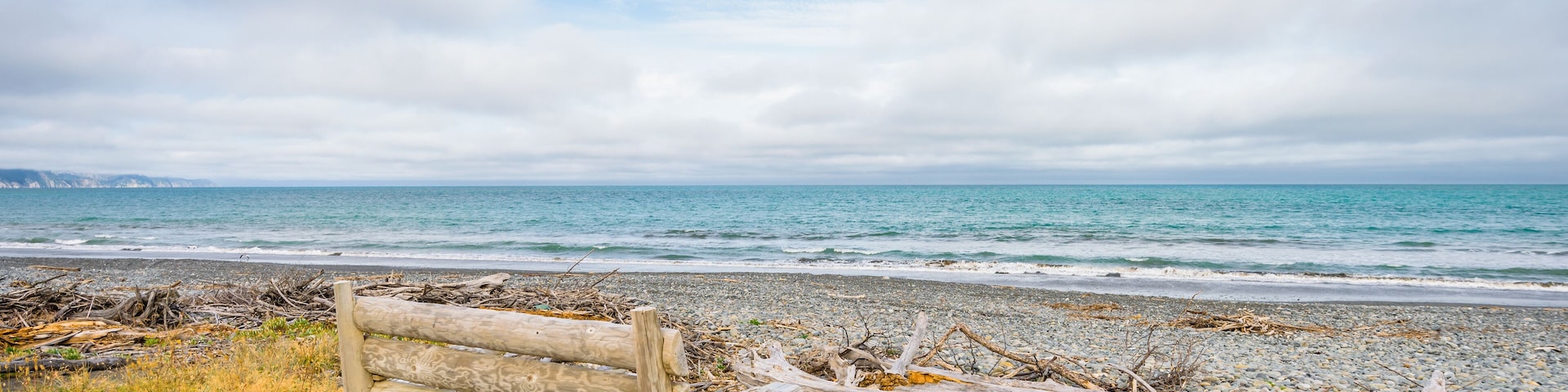 A wooden bench on a rocky beach with driftwood and a view of the ocean.