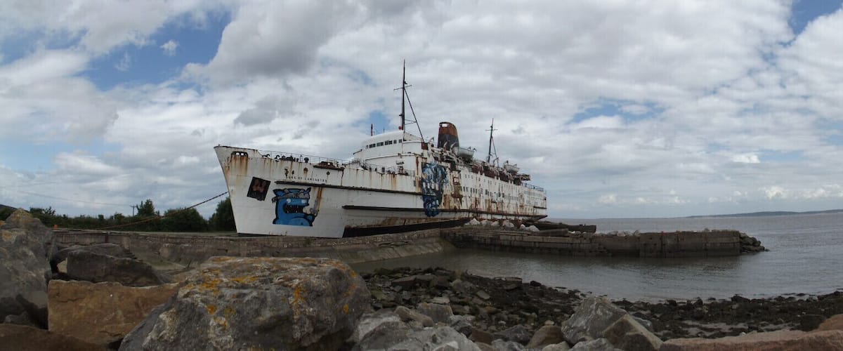 A rusted shipwreck on a rocky shore with a cloudy sky.