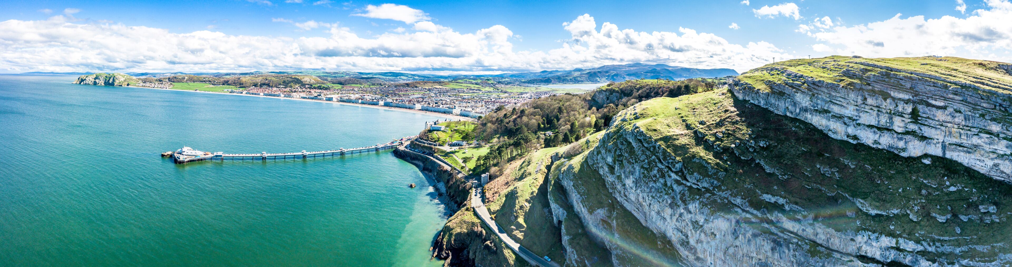A coastal town with a pier extending into the sea, a cliffside, and distant mountains.
