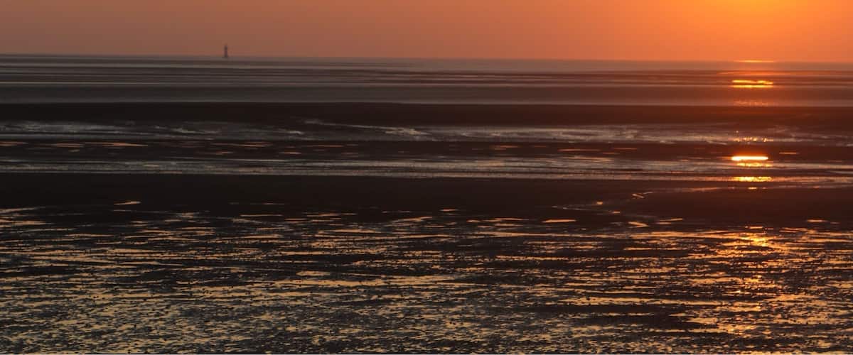 A sunset over a beach with waves and a distant lighthouse.