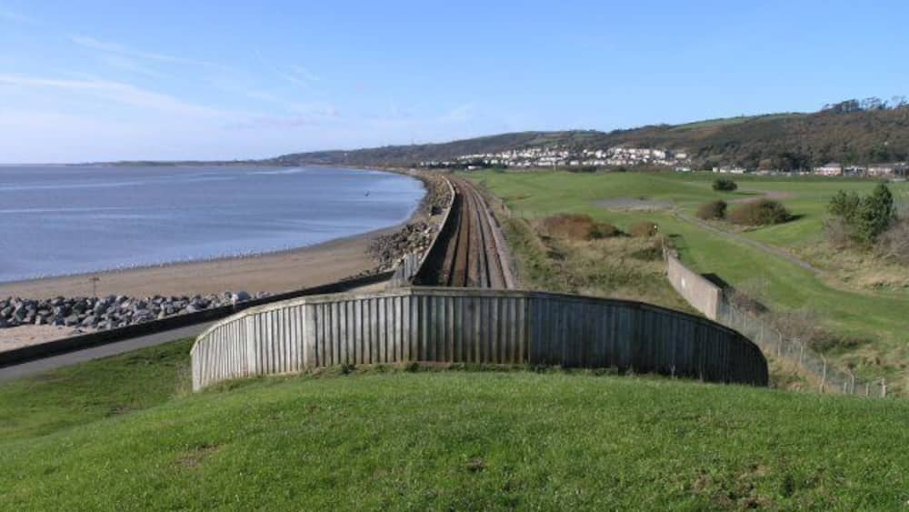 Eine Küstenstrecke der Eisenbahn schlängelt sich an einem Strand entlang, vorbei an grünen Feldern, mit einer Stadt in der Ferne.