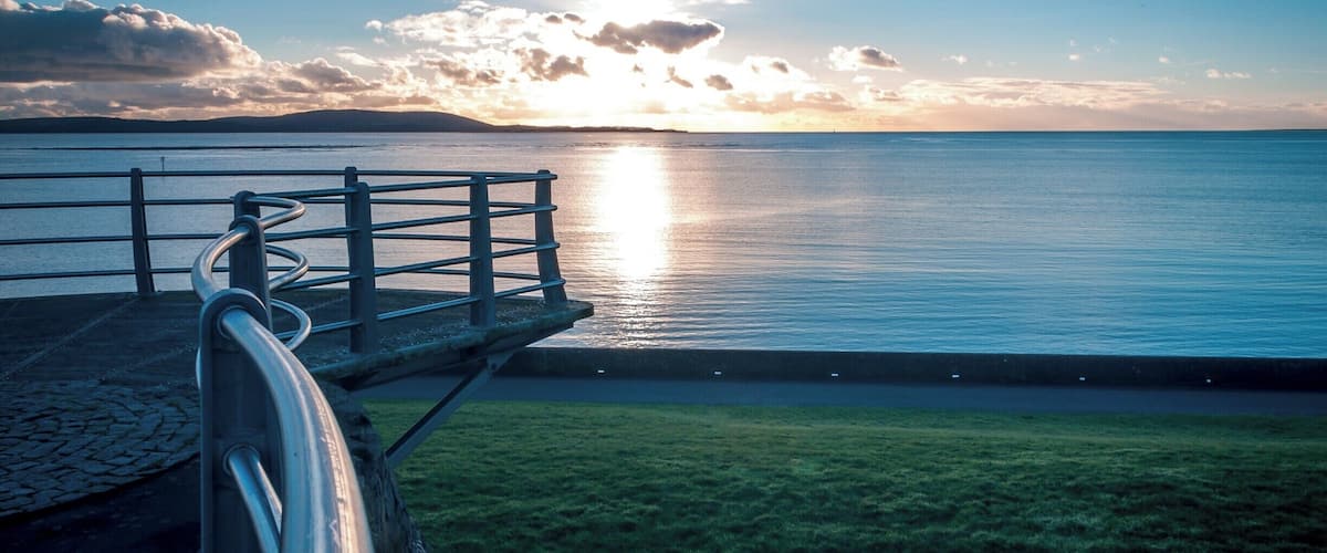 A waterfront promenade with a metal railing overlooking a calm sea and a sunset.