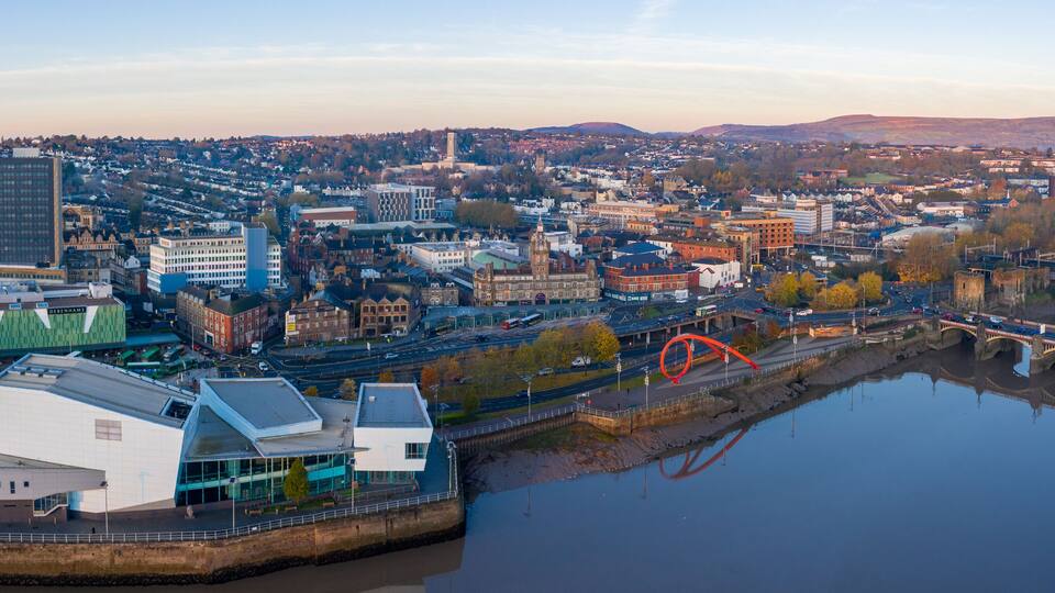 A cityscape with a river, a bridge, and various buildings including a modern structure and a historic tower.