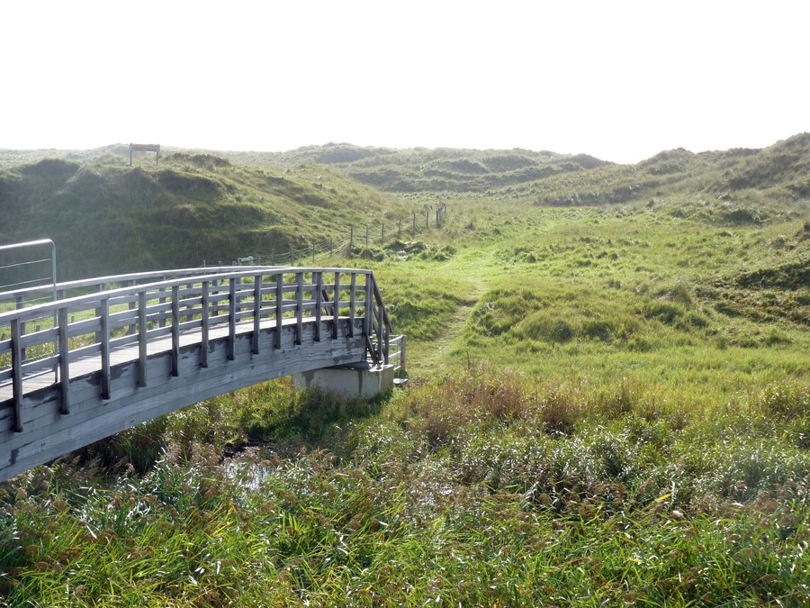 A wooden bridge over a grassy valley with hills in the background.