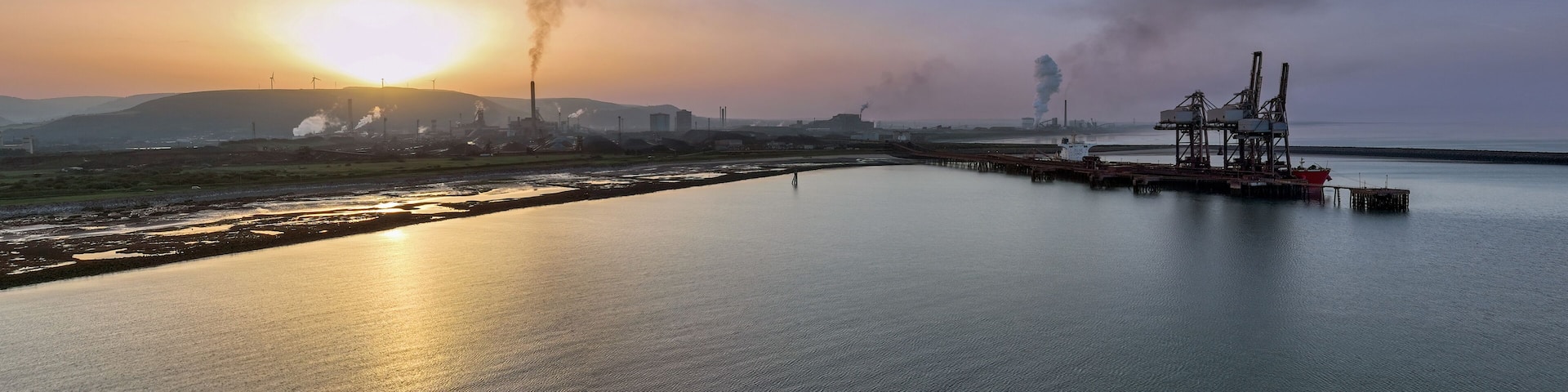A sunset over a calm body of water with industrial structures and smokestacks in the background.