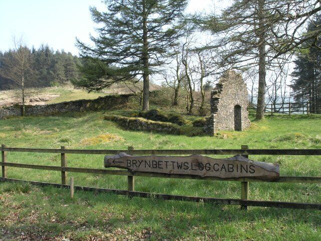 Un cartel de madera con la inscripción 'BRYNETTWS LOG CABINS', rodeado de una zona con césped, árboles y una estructura de piedra al fondo.