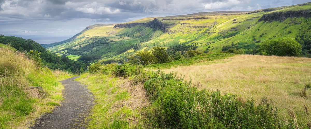 A winding path through a lush valley with rolling hills and a cloudy sky.