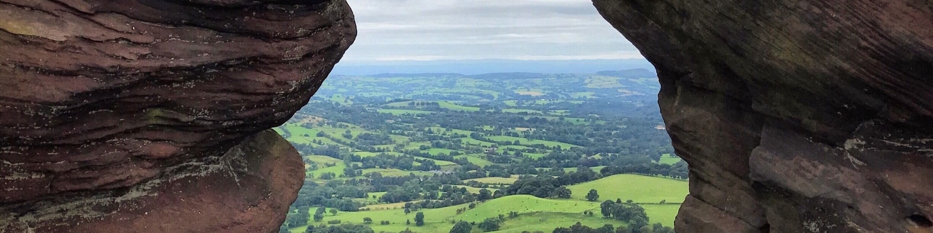 Paisagem com grandes formações rochosas emoldurando colinas verdes e montanhas distantes sob um céu nublado.