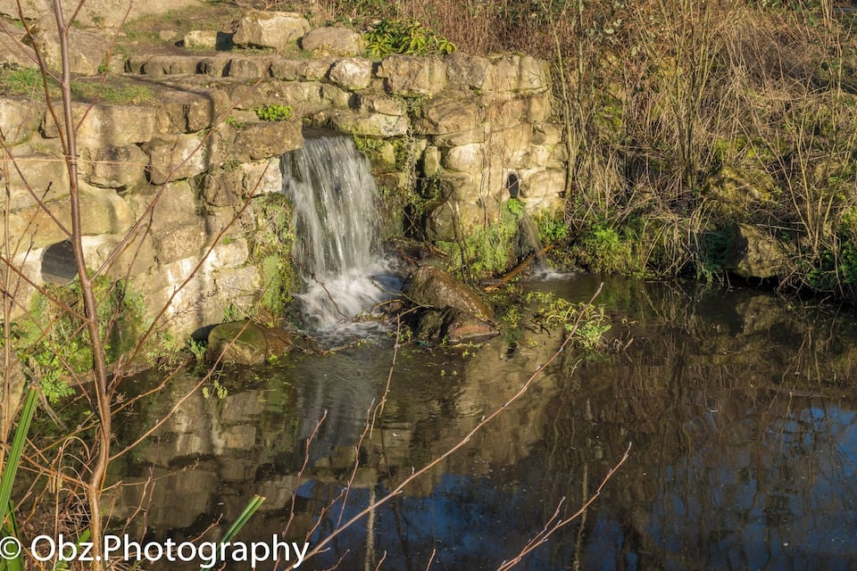 A small waterfall cascading over a stone wall into a pond surrounded by vegetation.