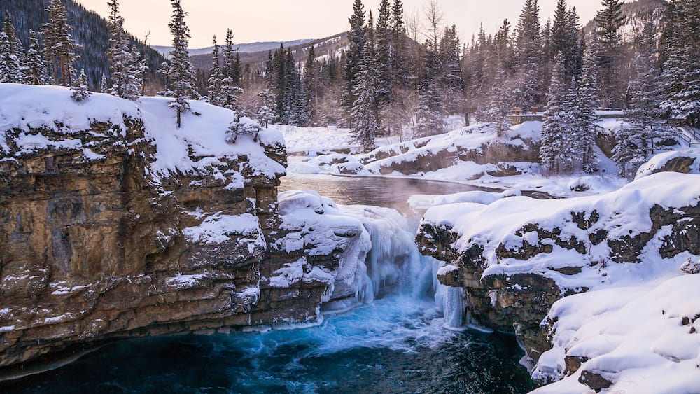 A snow-covered waterfall in a forested valley with rocky cliffs.
